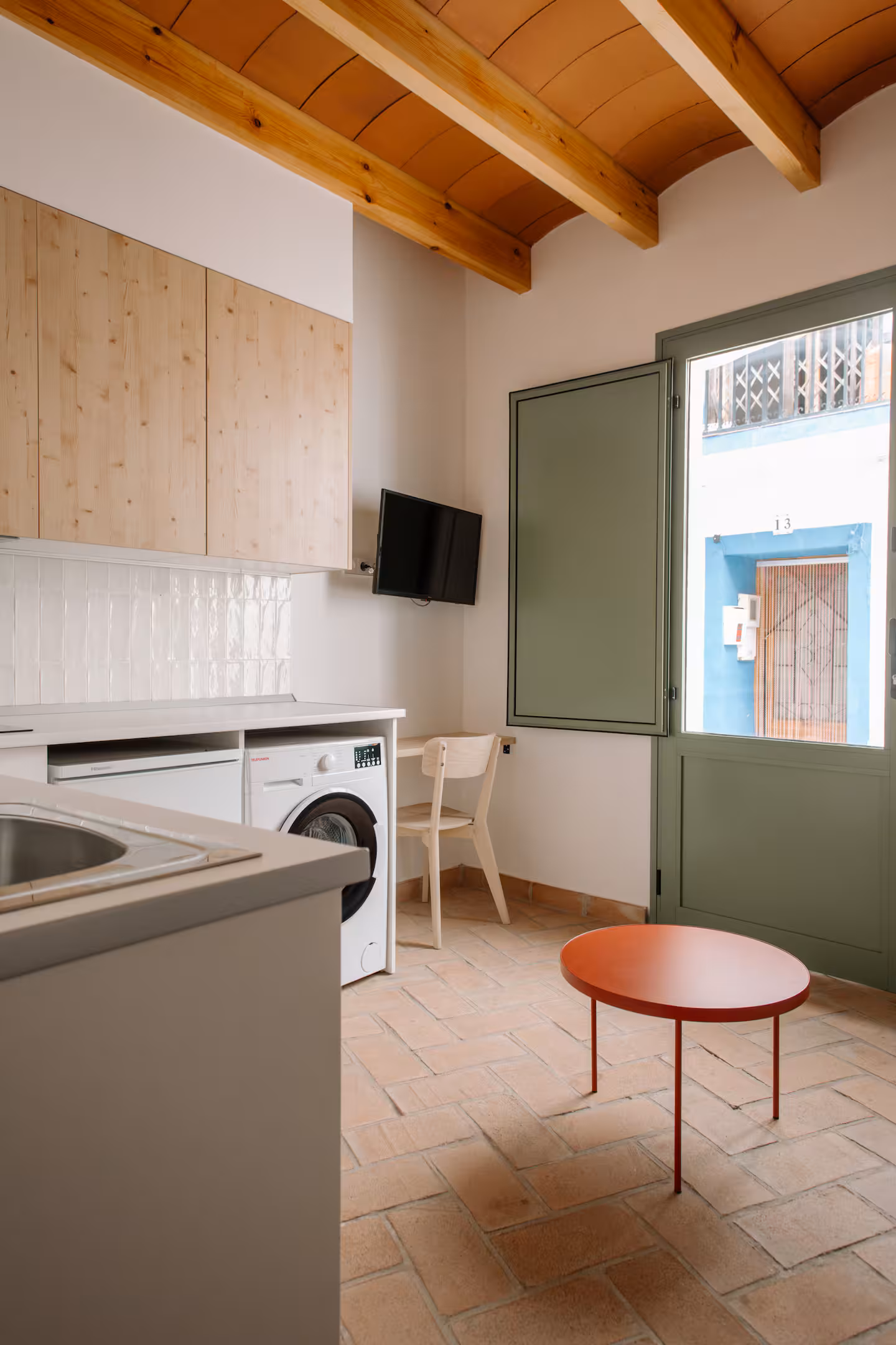 Cocina con contraventanas verdes abiertas a la calle, vigas de madera y mesa redonda roja