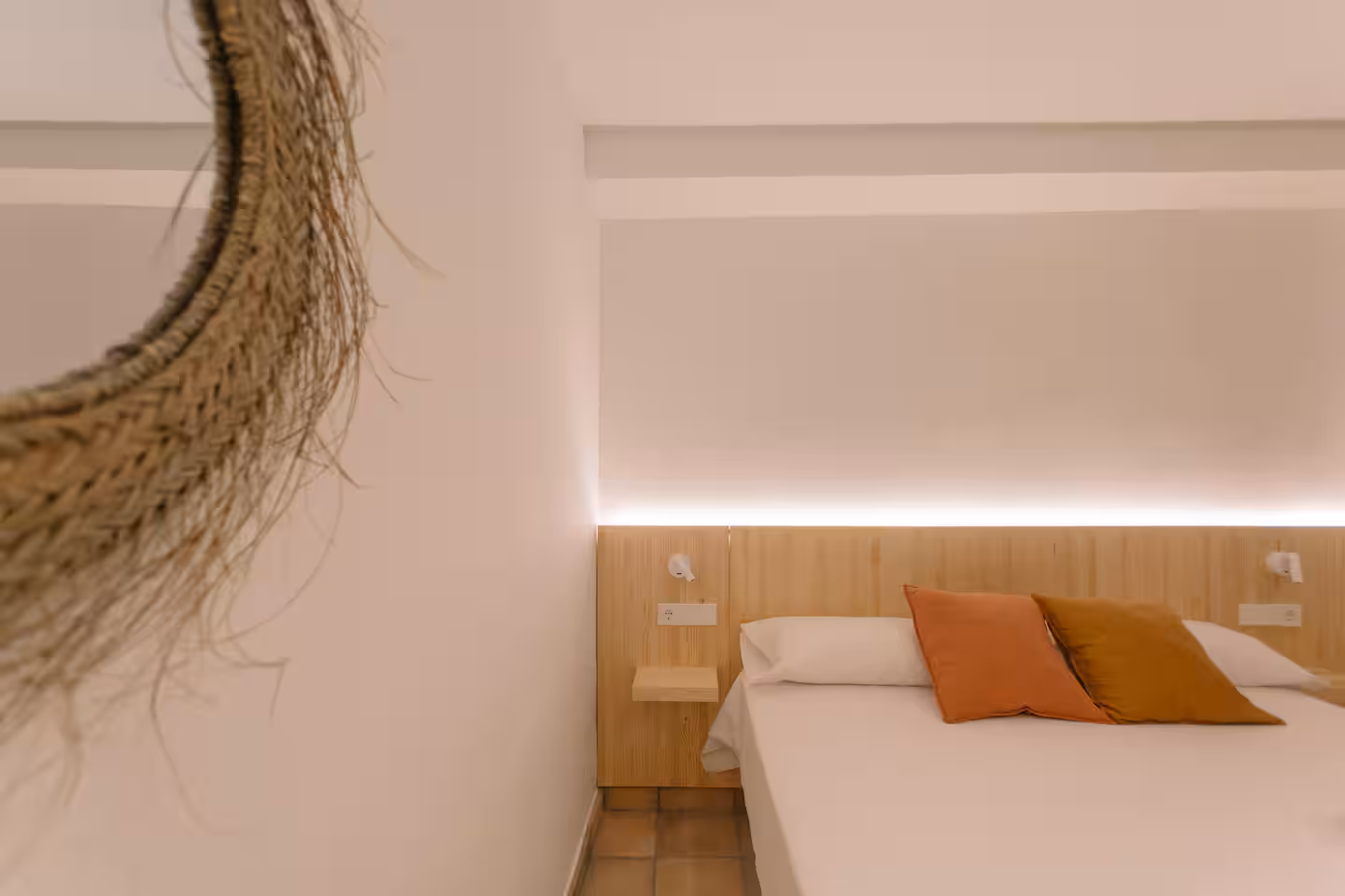 Cama vestida en blanco con cojines naranjas, luz indirecta en el cabecero y espejo de fibra