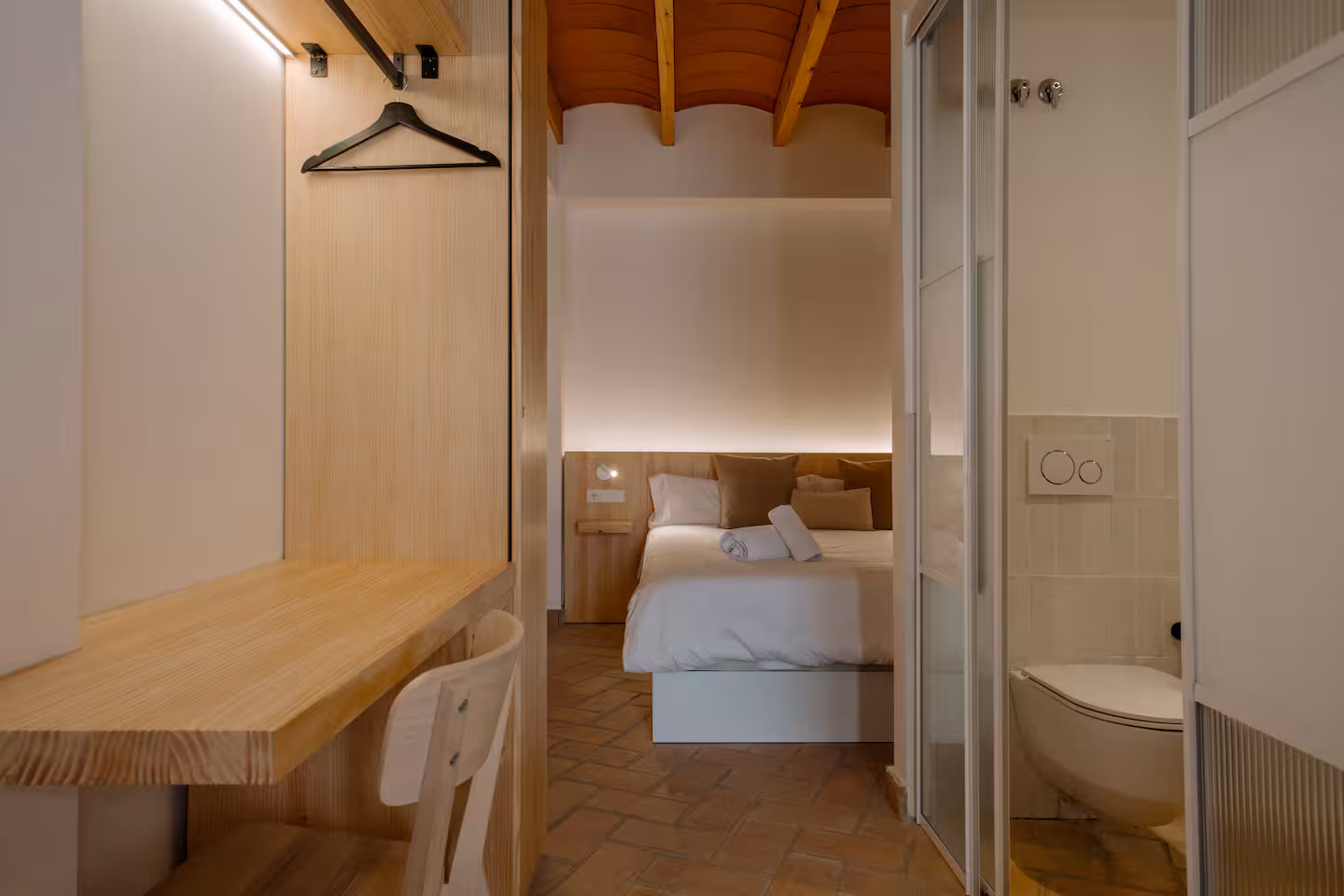 Perchero de madera, cama y acceso al baño bajo el techo de vigas